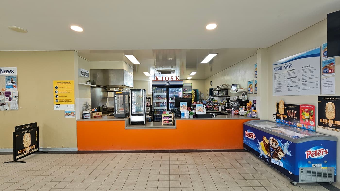The kiosk showcases a low orange reception counter with food racks and items. Behind it, a fridge stores cold drinks, and display racks hold snacks. To the right, an ice cream freezer is positioned. Advert posters and facility reminders are affixed to the walls, with an advert poster standing on the left.