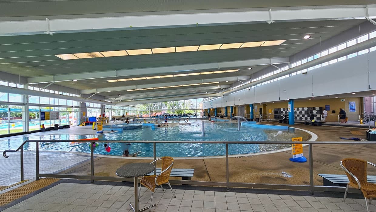 Next to the Leisure Pool is an elevated area near the cafe with movable tables and chairs. On the right, steps with handrails and truncated domes are present, along with bench chairs. The Leisure Pool is in front, and further to the right is the locker area. The upper wall is glass, offering an outside view with a large poster. The high ceiling includes strips of transparent roofing for natural sunlight and large aircon vents.