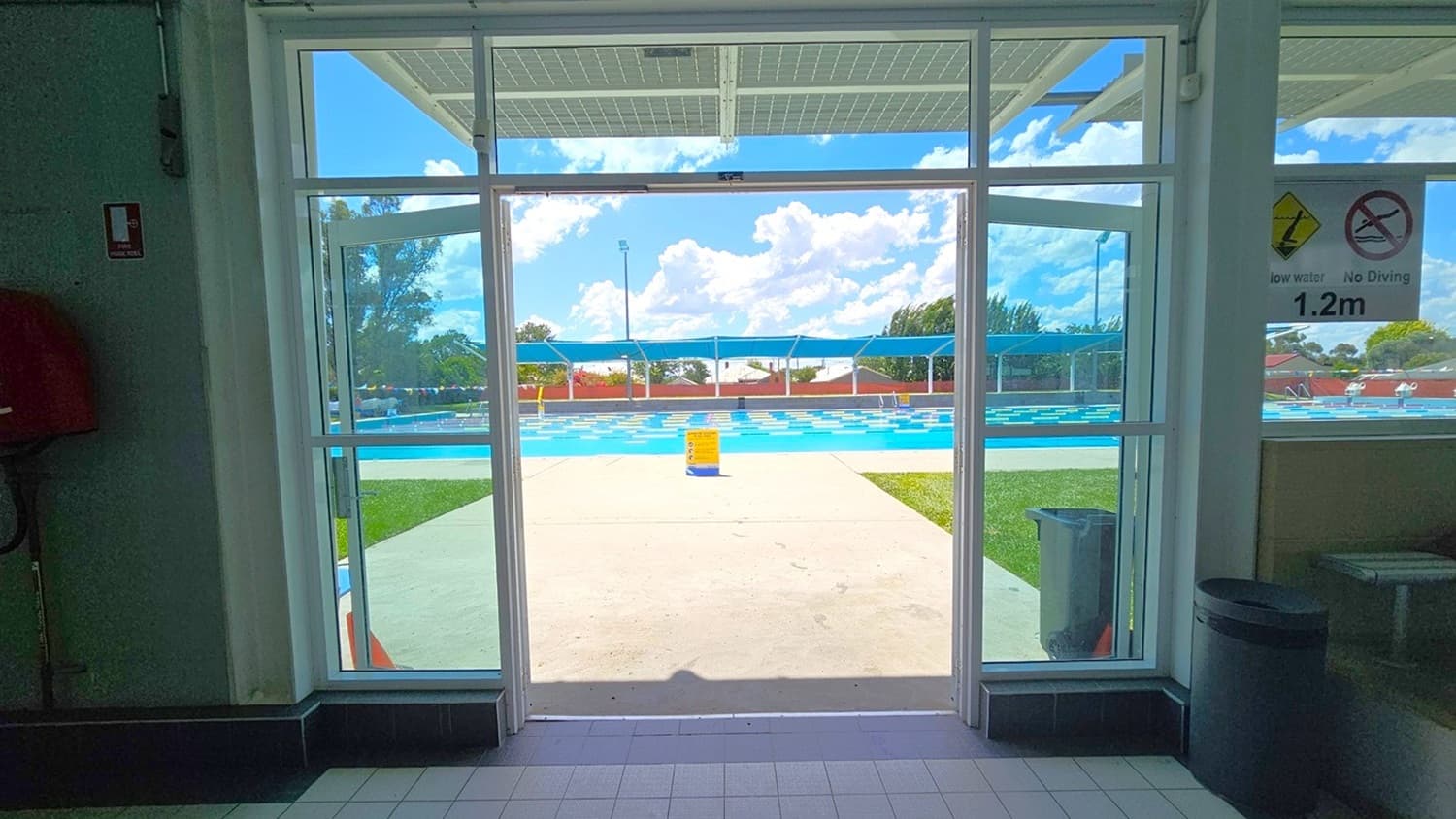 Looking through a glass door, a swimming pool is visible outside. The pool area has a tiled path leading to it, with grass on the right. A person is sitting by the pool, and there are blue shade structures above. Signs on the door indicate "1.2m" depth and "No Diving". Inside, the floor is tiled, and a rubbish bin is on the right. The ceiling inside is not visible.