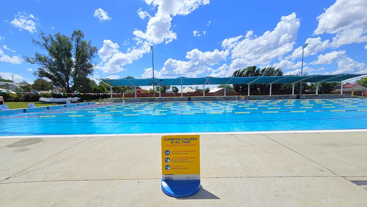 A large outdoor swimming pool with clear blue water is in the centre. In the foreground, a yellow sign on the pool deck advises supervising children. The pool is divided into lanes with floating markers. In the background, there are blue shade structures and a few trees. The ground is paved with light-coloured concrete.