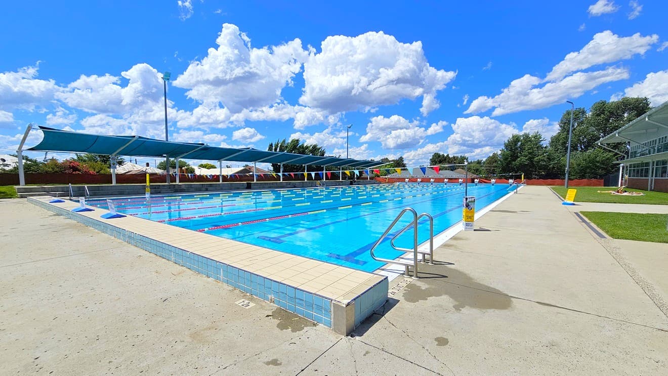 Outdoor swimming pool with clear blue water, surrounded by a concrete deck. On the left, there are several shaded areas with blue canopies. A metal handrail descends into the pool. On the right, a grassy area and a building with large windows are visible. The pool is divided into lanes with red and white lane markers. In the background, trees and a fence line the perimeter. The sky is filled with fluffy white clouds.