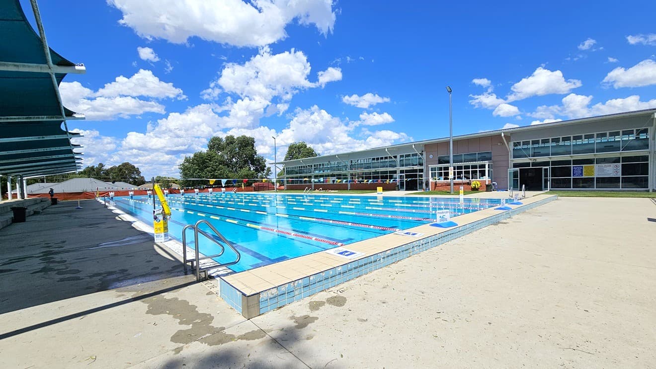 An outdoor swimming pool with clear blue water and lane dividers. On the left, there are shaded seating areas with dark blue canopies. The right side features a modern building with large glass windows. In the background, trees and a few clouds are visible. The pool deck is light-coloured concrete with a metal handrail leading into the water.