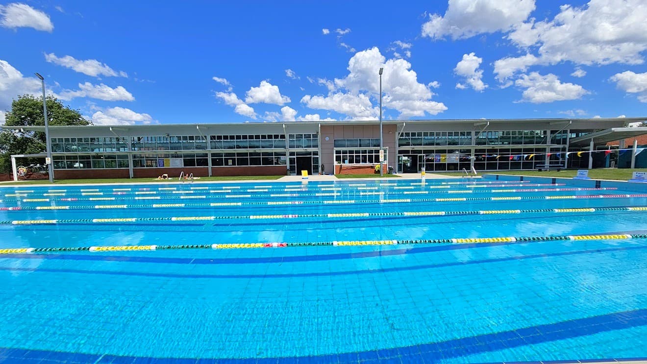 An outdoor swimming pool with multiple lanes marked by yellow and black lane dividers. In the background, a modern building with large glass windows stretches across the image. A few people are visible near the building, possibly sunbathing or resting. The pool area is surrounded by a grassy lawn, and there are tall lights on poles. The water in the pool is clear and blue, reflecting the surroundings.