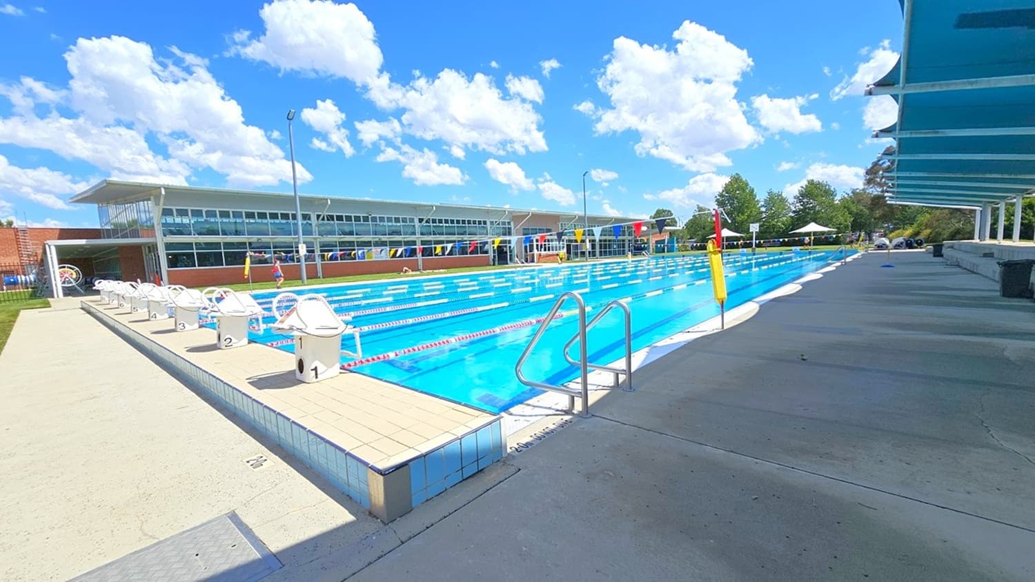 An outdoor swimming pool with multiple lanes, surrounded by a concrete deck. On the left, there are starting blocks numbered sequentially. A modern building with large windows is in the background. On the right, a covered walkway provides shade. Trees and a small tent are visible in the distance. The pool is equipped with lane dividers and a metal ladder for entry.