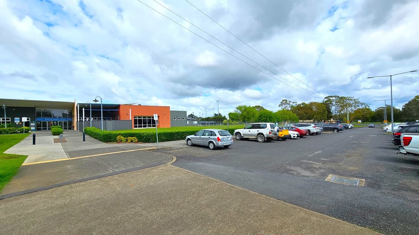 A modern building with large glass windows is on the right, labelled "Aqua Centre" and "Aqua Aerobics". The structure has a slanted roof and is surrounded by a well-maintained grassy area. On the left, there are several trees and a wooden fence running parallel to a concrete path. The path continues on the right, lined with street lamps and more trees. The sky is overcast.