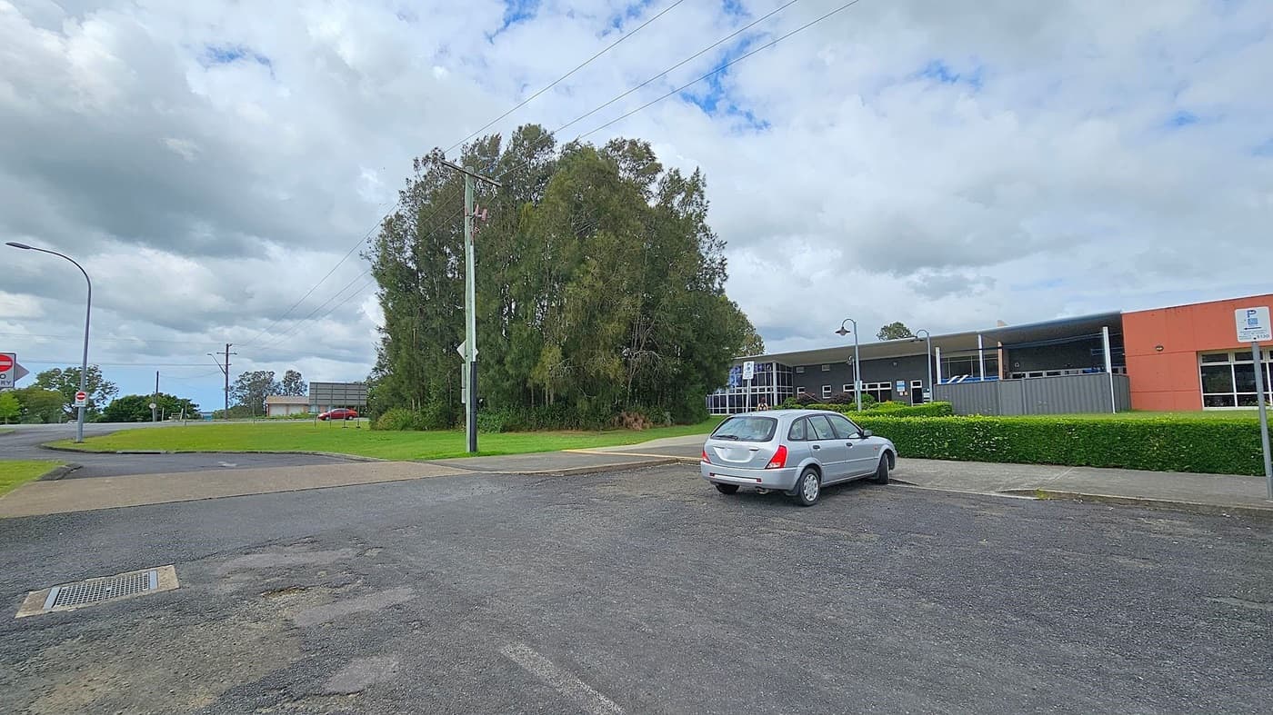 A silver car is parked on a paved area in the foreground. To the right, there's a building with a red and grey facade, surrounded by a neatly trimmed hedge. In the background, a cluster of tall trees stands near a grassy area. A red car is visible in the distance on a road. A streetlamp and a signpost are on the left, with a cloudy sky above.