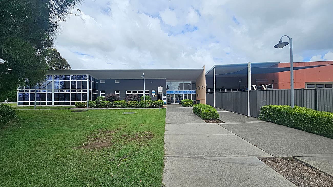 A modern building with a flat roof and large glass windows on the left side. The entrance is in the centre, with a sign above the doors. The right side features a metal fence and a covered walkway. The foreground has a concrete path flanked by green grass and neatly trimmed bushes. A streetlamp is visible on the right.