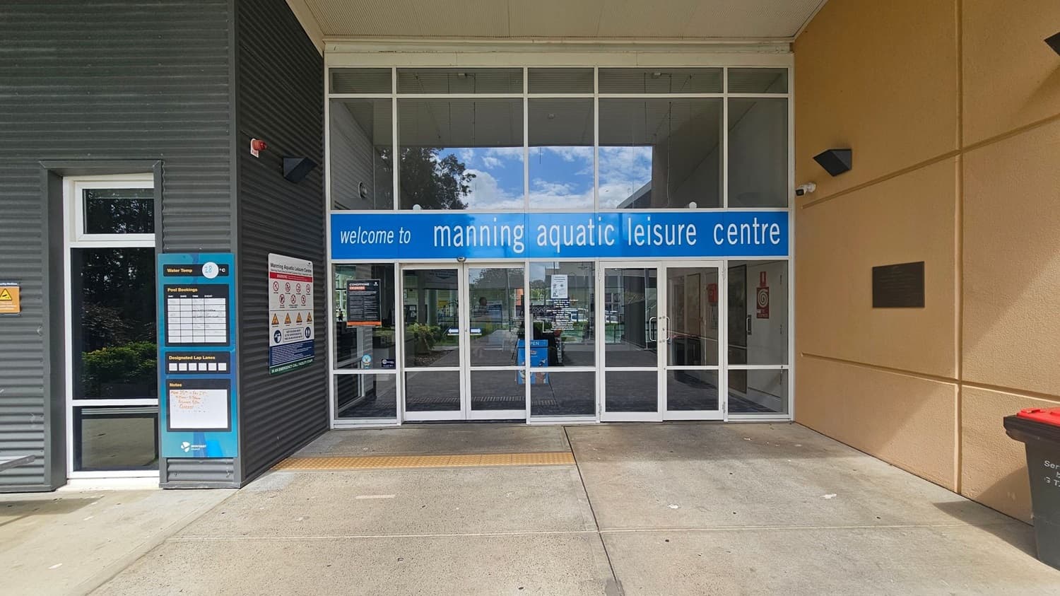 Entrance to the Manning Aquatic Leisure Centre with a large glass door in the centre. Above the door is a blue sign with white text. On the left, there are information boards displaying water temperature and pool bookings. The wall is dark grey with a small window. On the right, the wall is beige with a black rectangular fixture and a red-lidded bin. The floor is concrete, and the ceiling is light-coloured.