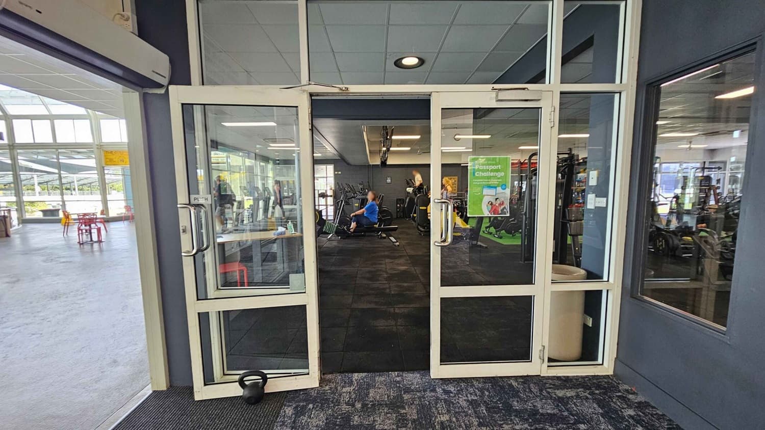 Glass doors open into a gym area with exercise equipment visible inside. A person in a blue shirt is using a rowing machine. To the left, there is an open space with red chairs and tables. A poster on the door advertises a "Passport Challenge." The floor inside the gym is dark, while the area outside has a lighter, speckled carpet. The ceiling is fitted with recessed lighting.