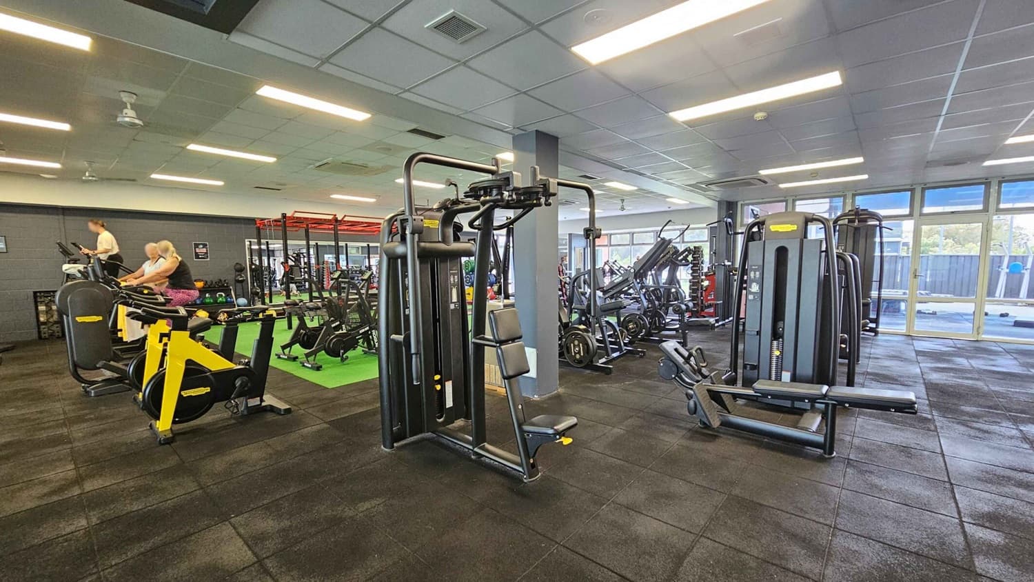 A gym interior with various exercise equipment. On the left, there are stationary bikes and a treadmill with people using them. In the centre, a multi-functional weight machine is visible. To the right, more weight machines and benches are arranged. The floor is covered with dark rubber tiles, and the ceiling has fluorescent lights. Large windows in the background allow natural light to enter.