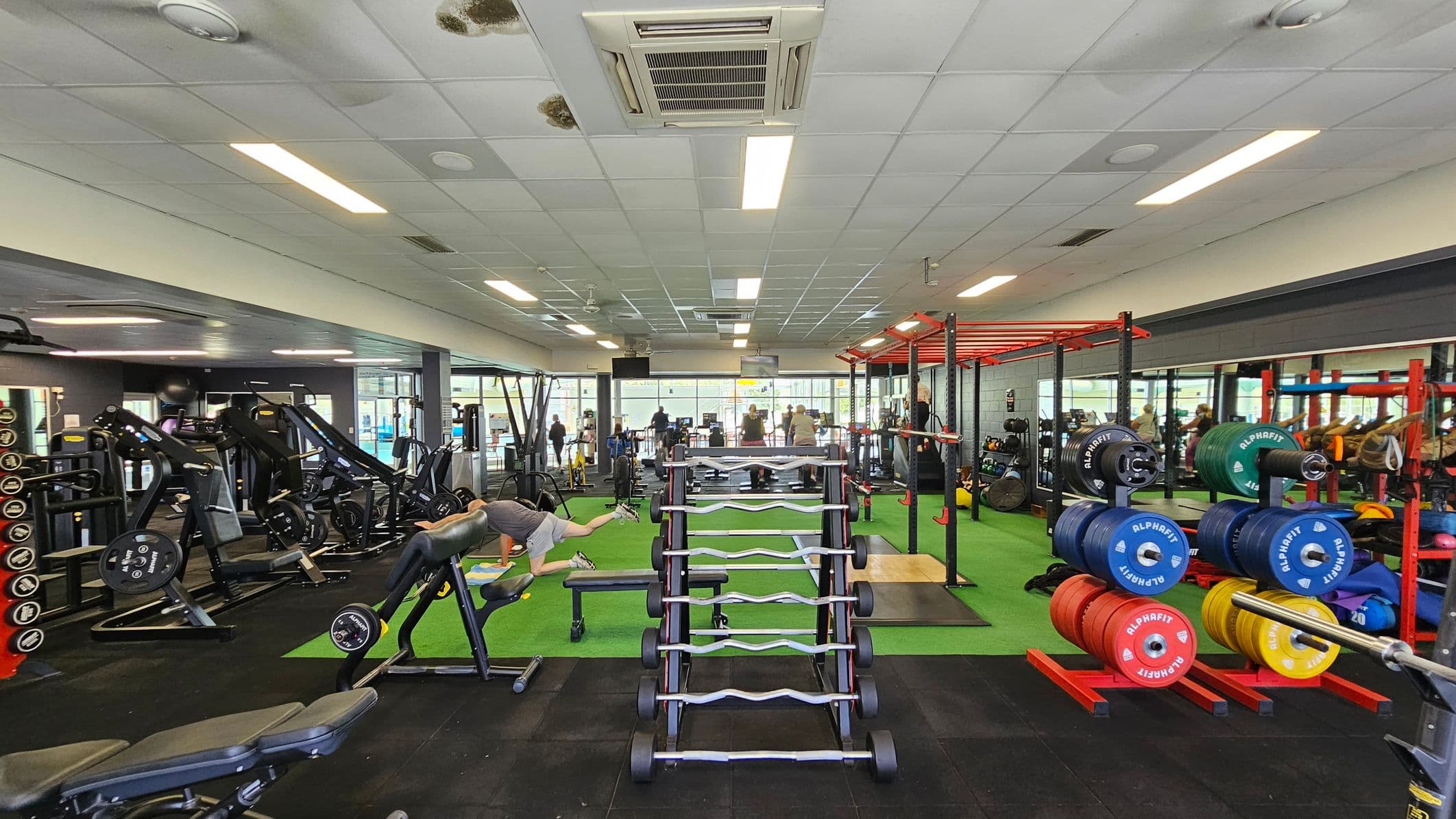 A gym interior with various exercise equipment. On the left, there are weight machines and benches. In the centre, a rack of barbells is visible, with a person exercising nearby. On the right, colourful weight plates are stacked on racks, and a red metal frame structure is present. The floor is a mix of black and green surfaces, and the ceiling has fluorescent lights and air conditioning vents. In the background, people are using treadmills near large windows.