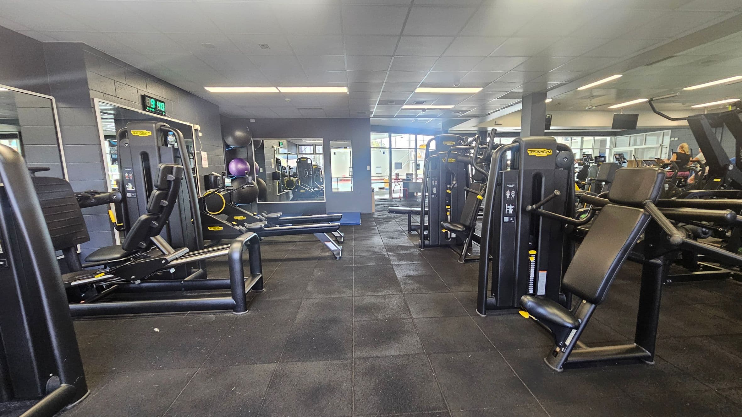 A gym interior with various exercise machines on both sides. On the left, there are weight machines and a wall-mounted digital clock displaying 9:40. The right side features more workout equipment, including a seated machine with a padded backrest. In the background, there are large windows allowing natural light, and a few exercise balls are visible. The floor is covered with black rubber tiles, and the ceiling has a grid pattern with fluorescent lights.