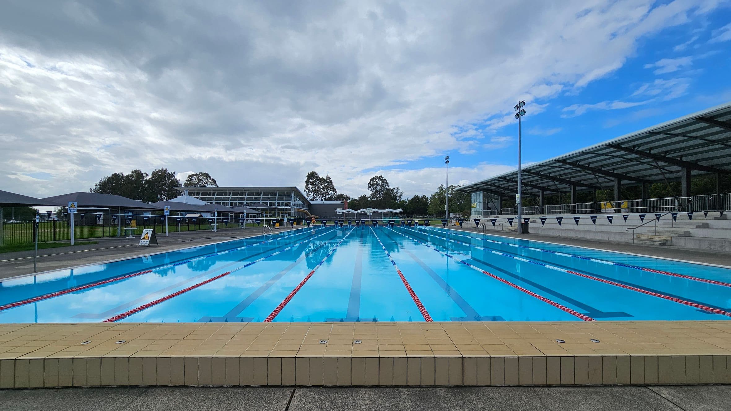 An outdoor swimming pool with clear blue water and red lane dividers stretches into the distance. On the right, there is a covered seating area with a metal railing and steps. On the left, a grassy area with a fence and several small structures is visible. In the background, there are trees and a large building with a glass facade. The pool deck is made of concrete, and the pool edge is lined with yellow tiles.