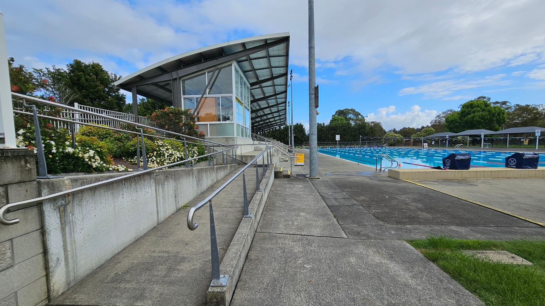 On the left, there is a modern building with a sloped roof and large glass windows. A concrete ramp with metal railings leads up to it, bordered by a garden with white and red flowers. On the right, there is a large outdoor swimming pool with blue water, divided into lanes. The pool area is surrounded by a paved walkway and some grassy patches. In the background, there are trees and a few covered seating areas.
