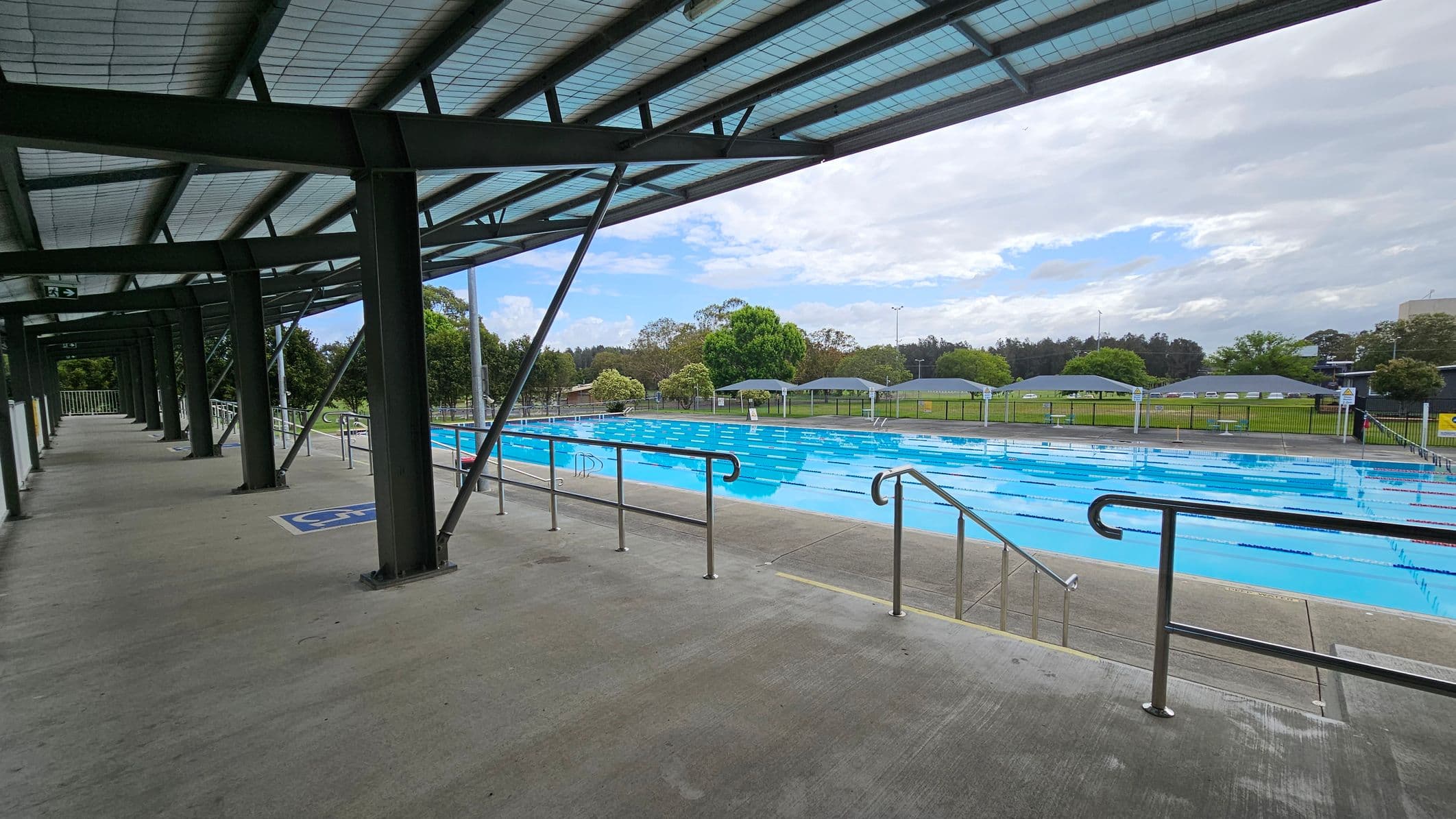 A covered walkway on the left with a concrete floor and metal beams supporting a translucent roof. On the right, an outdoor swimming pool with clear blue water and lane dividers. Beyond the pool, there are green lawns, trees, and several shaded seating areas. The background features more trees and a fence enclosing the area.