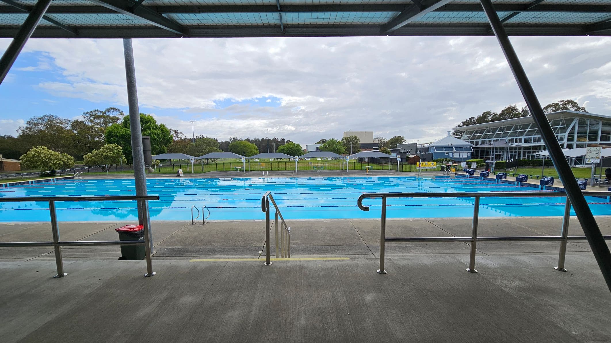 An outdoor swimming pool with clear blue water is in the centre, surrounded by a concrete deck. Metal railings and steps lead into the pool. On the right, a modern building with large glass windows is visible. In the background, there are green trees and a grassy area with small shelters. The ceiling is a metal structure with beams. A rubbish bin is placed near the railing.