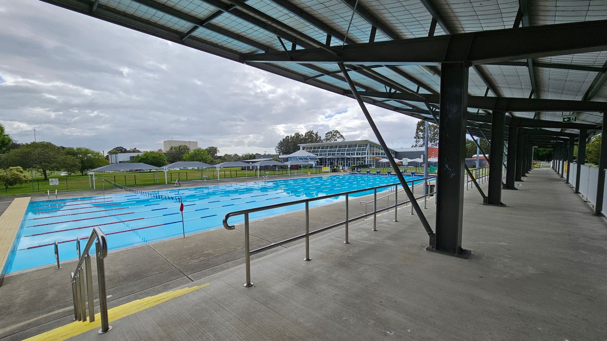 A covered walkway with a metal roof and support beams runs along the right side. On the left, there is a large outdoor swimming pool with lane dividers. Beyond the pool, there are green lawns and trees, with a modern glass building in the background. The floor is concrete, and there are metal railings along the walkway.
