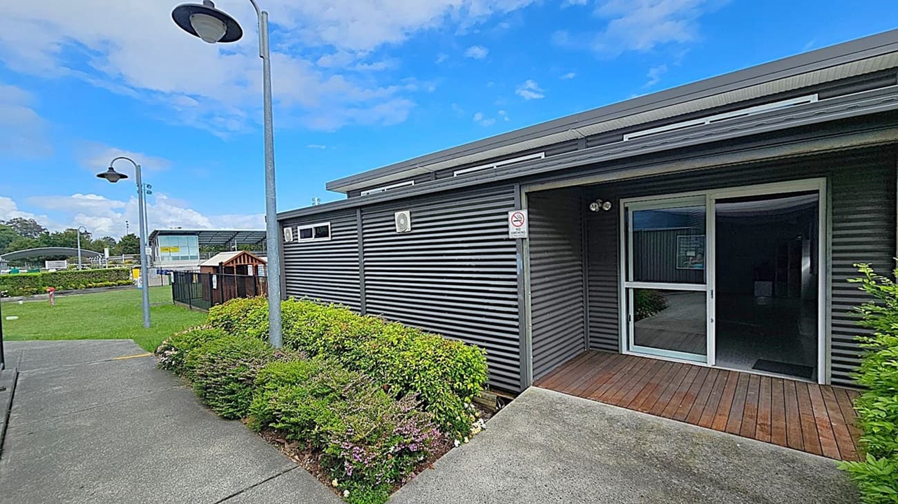 A small entrance with a wooden deck leads to a building with corrugated metal walls. A glass sliding door is in the centre, partially open, revealing a dimly lit interior. A "No Smoking" sign is mounted on the left wall. On the right, there is a lush green bush next to a white metal fence. The roof is flat with a metal overhang. In the background, there is a grassy area with trees and a road.