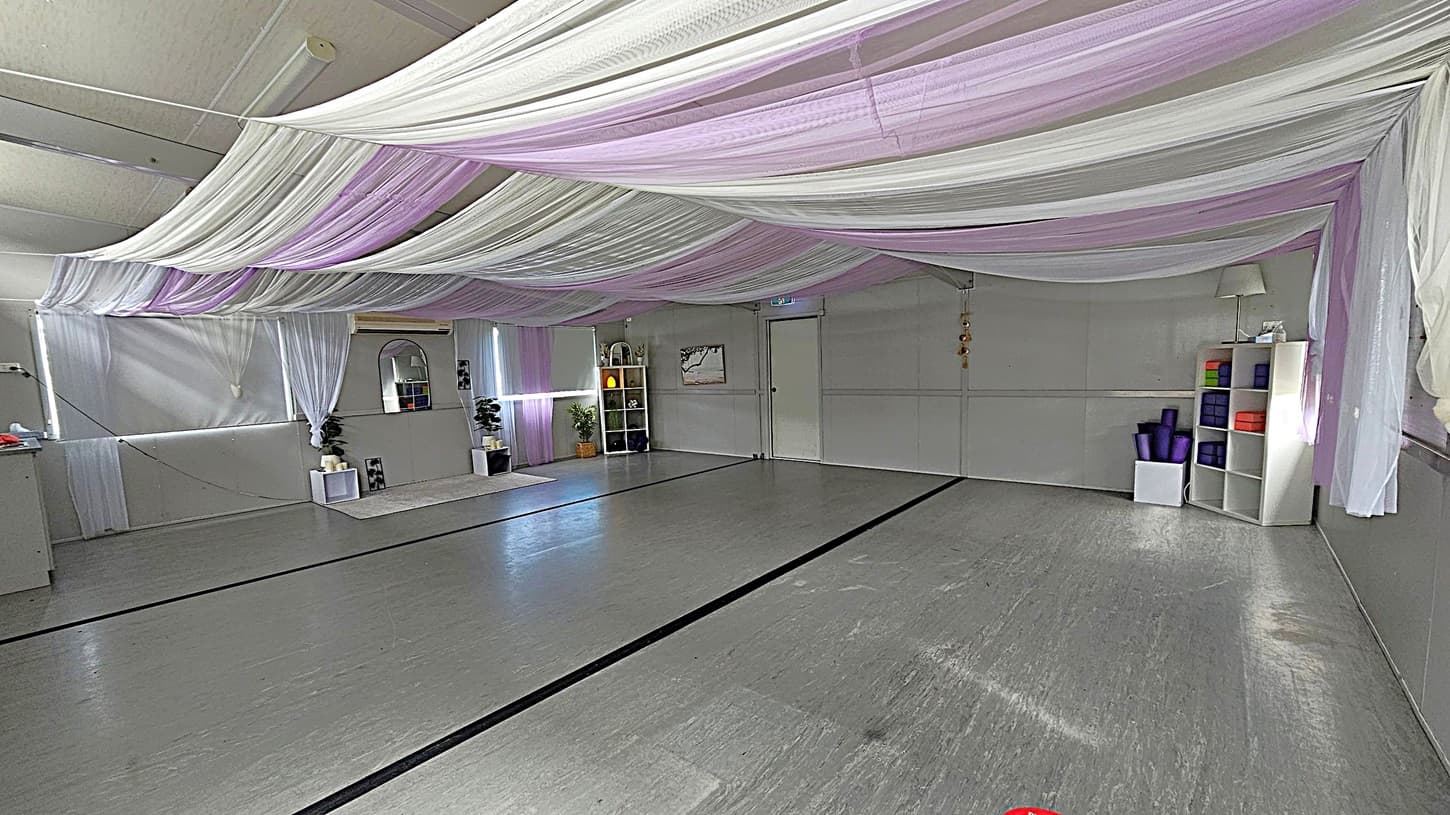 A spacious room with a grey floor and ceiling draped with white and lavender fabric. On the left, there are white shelves with potted plants and a mirror. In the centre, a white door is visible. On the right, a white shelving unit holds yoga mats and blocks. The walls are light grey, and the room is softly lit, creating a calm atmosphere.