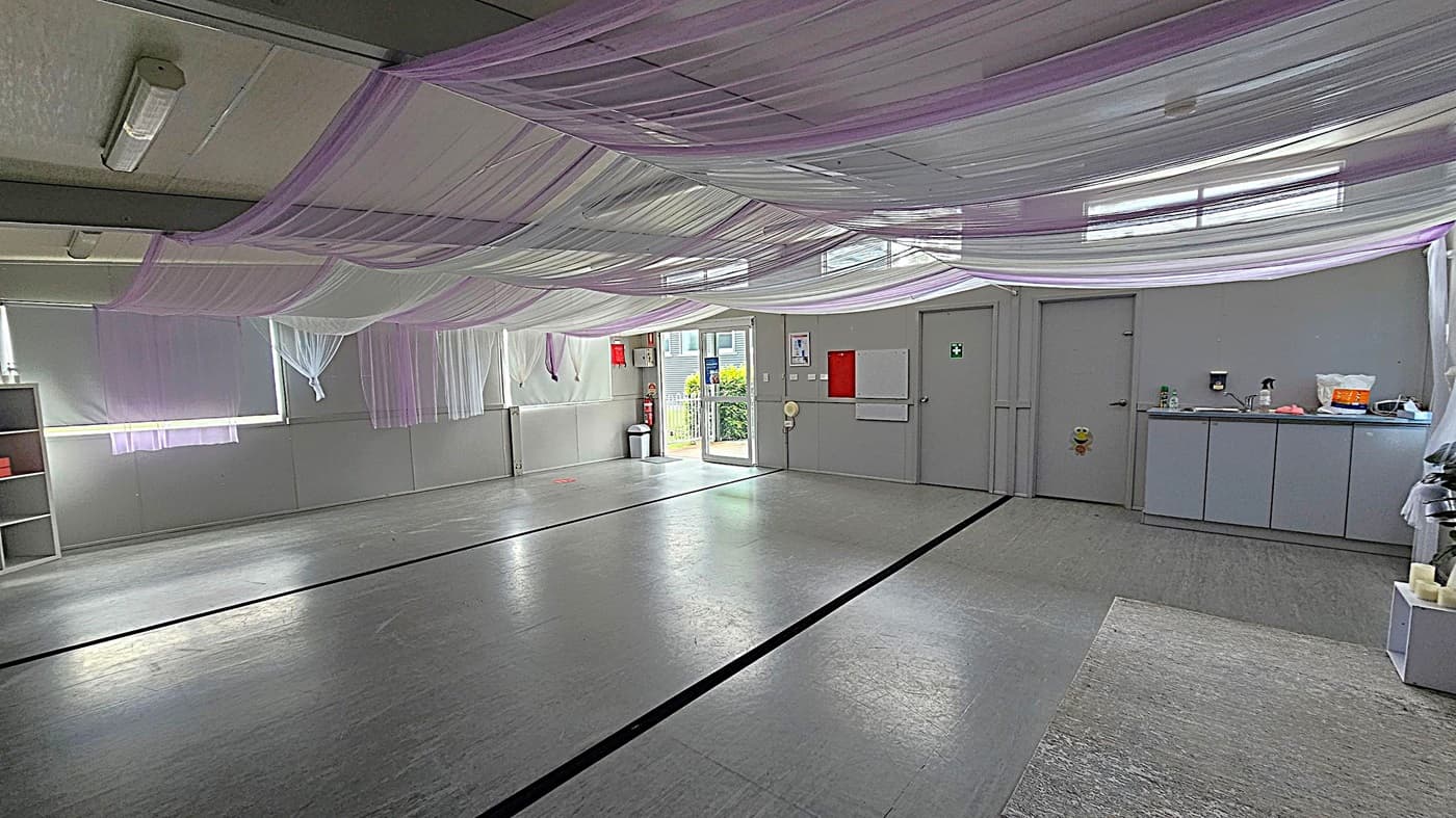A spacious room with a grey floor and ceiling, adorned with draped white and pink fabric. On the left, there are large windows covered with sheer curtains and a small shelf. In the centre, a glass door leads outside, flanked by a fire extinguisher and a bin. On the right, there are two closed doors and a countertop with a sink, cleaning supplies, and a few items. The room is well-lit with fluorescent lights.