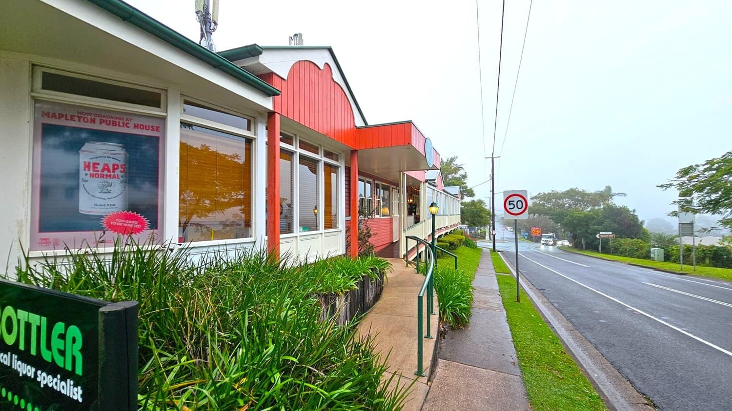 A street scene with a building on the left featuring large windows and a poster advertising a beverage. The building has a red and white facade with a green roof. In front, there is a path lined with green plants and a handrail. On the right, a road with a 50 km/h speed limit sign runs alongside the building. Vehicles are visible in the distance, and trees and greenery line the road. The sky is overcast, creating a misty atmosphere.