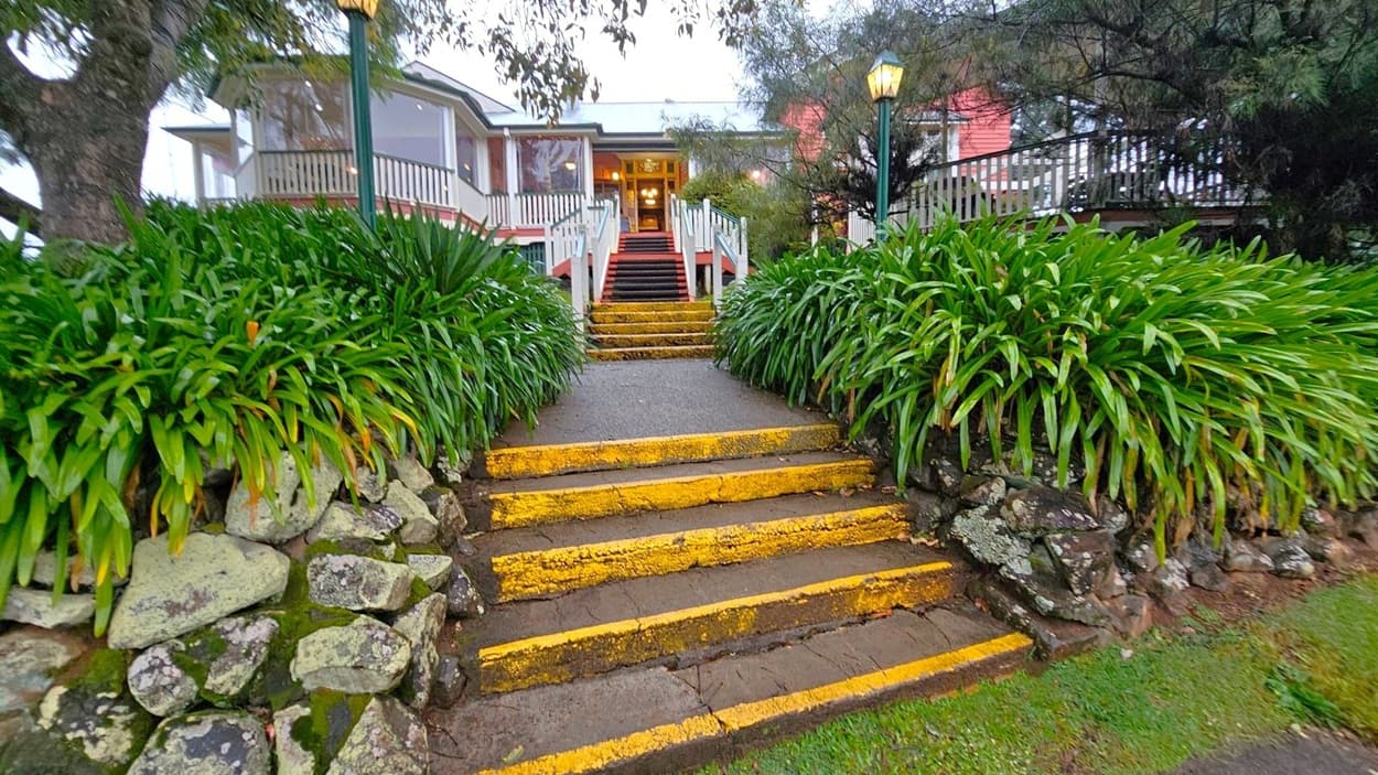 A pathway with yellow-edged steps leads up to a building with a porch and large windows. The steps are flanked by lush green plants and stone walls. Two lamp posts are visible on either side of the path. The building has a white and red exterior with a grey roof. Trees with dense foliage are on both sides, and the ground is covered with grass.
