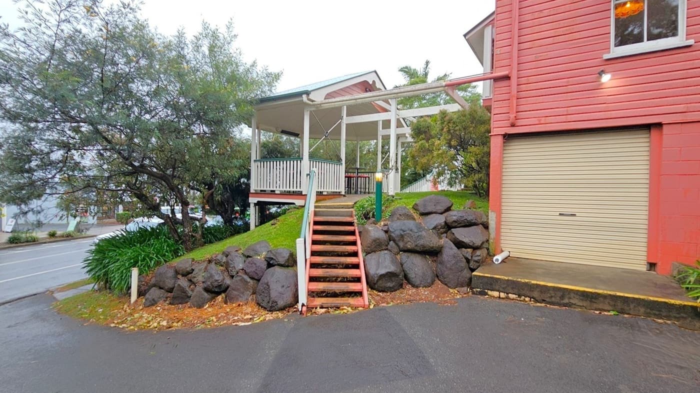 A small wooden structure with a white railing and a sloped roof is elevated on a grassy mound. Wooden steps with a handrail lead up to it, flanked by large dark rocks. To the right, a red building with horizontal siding features a closed beige garage door. A tree with dense foliage is on the left, near a road with visible markings. The ground is covered with fallen leaves, and a light fixture is mounted on the building.