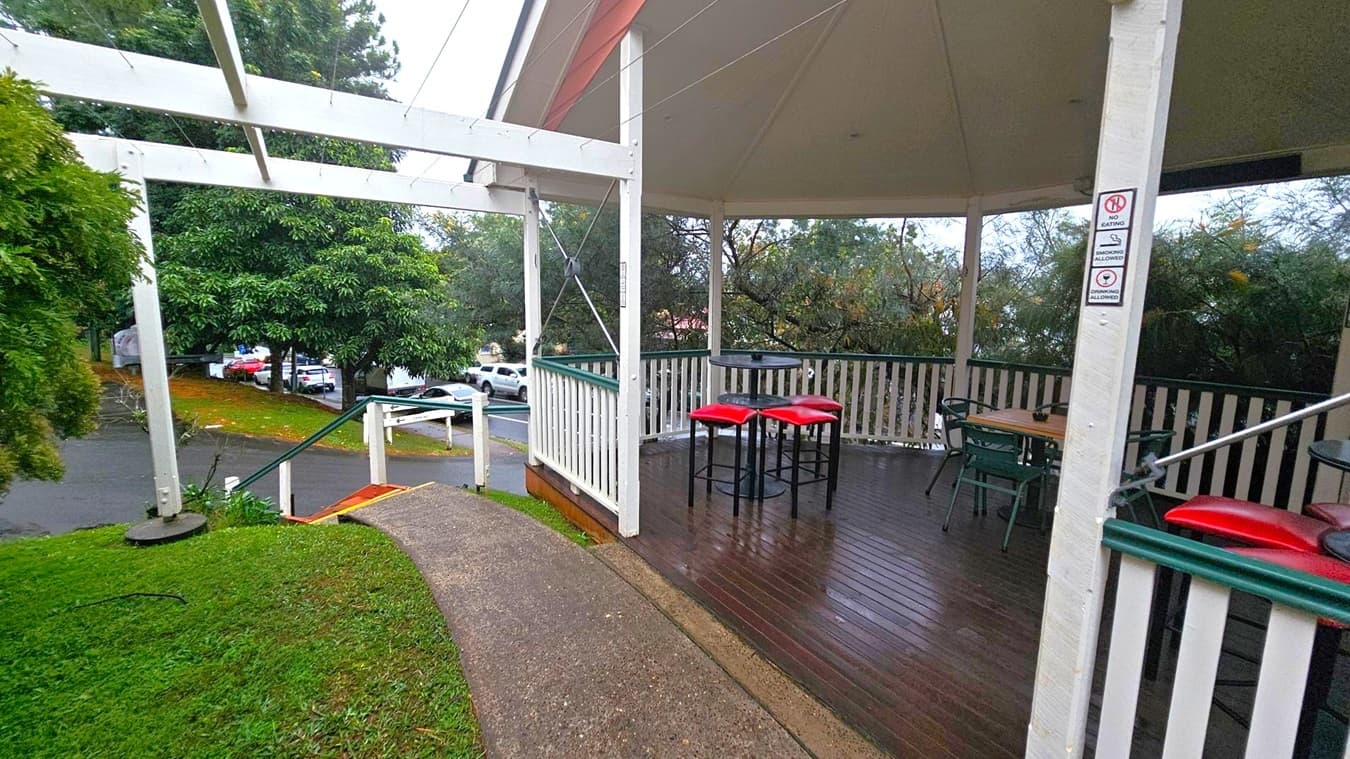 A covered outdoor seating area with a wooden deck and white railing. On the right, there are tables with red stools and chairs. Signs on a post indicate restrictions. The left side shows a sloped path leading downwards, bordered by grass and trees. In the background, a street with parked cars is visible. The structure has a white frame and a slanted roof.