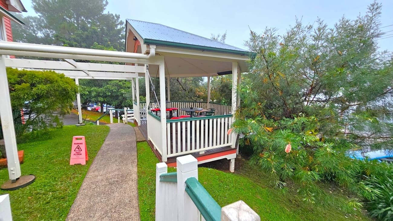 A small gazebo with a green roof and white railings is on the right, surrounded by dense greenery. Inside, there are tables and chairs. A concrete path leads to the gazebo, with a pink "wet floor" warning sign on the left. A white pergola structure is above the path. In the background, there are trees and a glimpse of parked cars. The ground is covered with grass.
