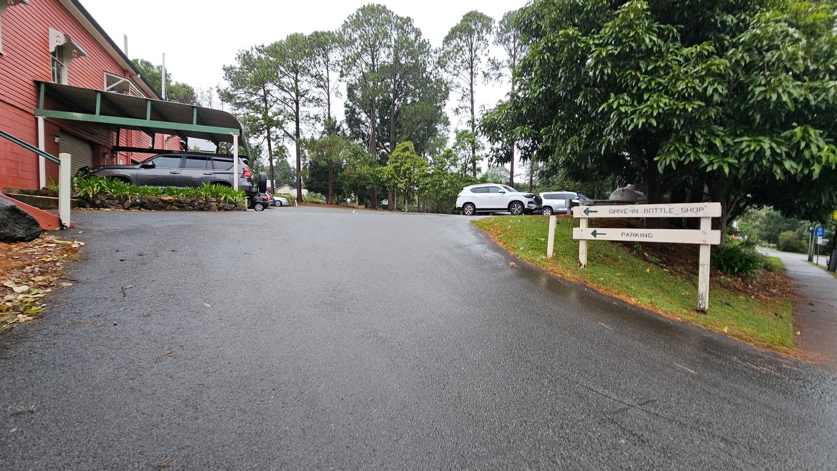 A sloped asphalt driveway leads up to a building on the left with a red brick exterior and a green awning. A grey car is parked under the awning. On the right, a grassy area with a large tree and a white car parked nearby. A wooden signpost indicates directions to a "Drive-In Bottle Shop" and "Parking." In the background, tall trees line the horizon, and additional cars are visible. The ground is slightly wet, suggesting recent rain.