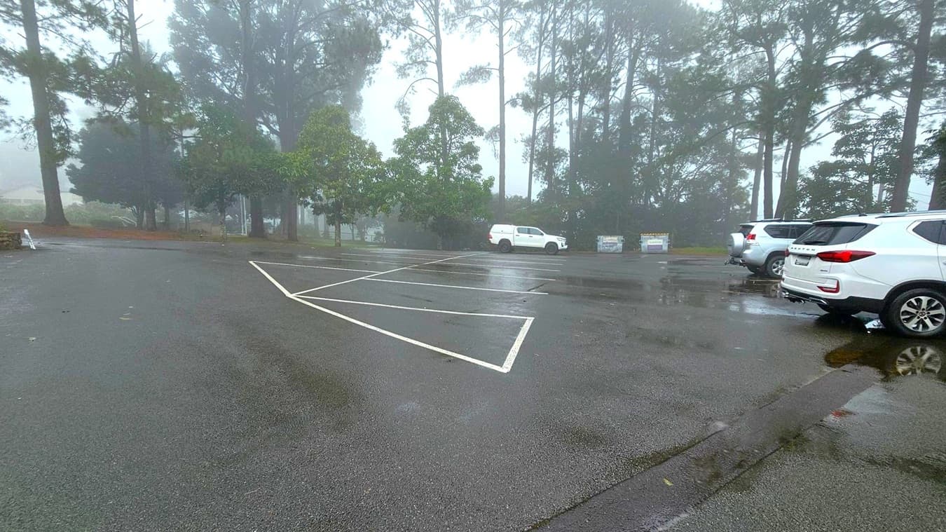 A misty car park with wet asphalt and visible puddles. On the right, several white SUVs are parked. In the background, tall trees are partially obscured by fog, creating a dense forested area. The left side shows empty parking spaces with white lines. The atmosphere is damp and overcast, with reflections of the trees visible in the water on the ground.
