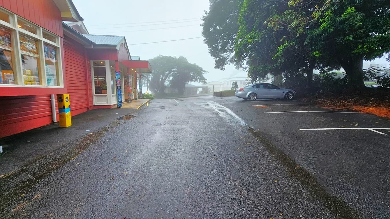 A misty street scene with a wet asphalt road. On the left, there is a red building with large windows and a covered entrance. A yellow bin is near the building. On the right, a silver car is parked under large, leafy trees. The background is foggy, obscuring distant buildings and trees. The road has some puddles and scattered leaves.