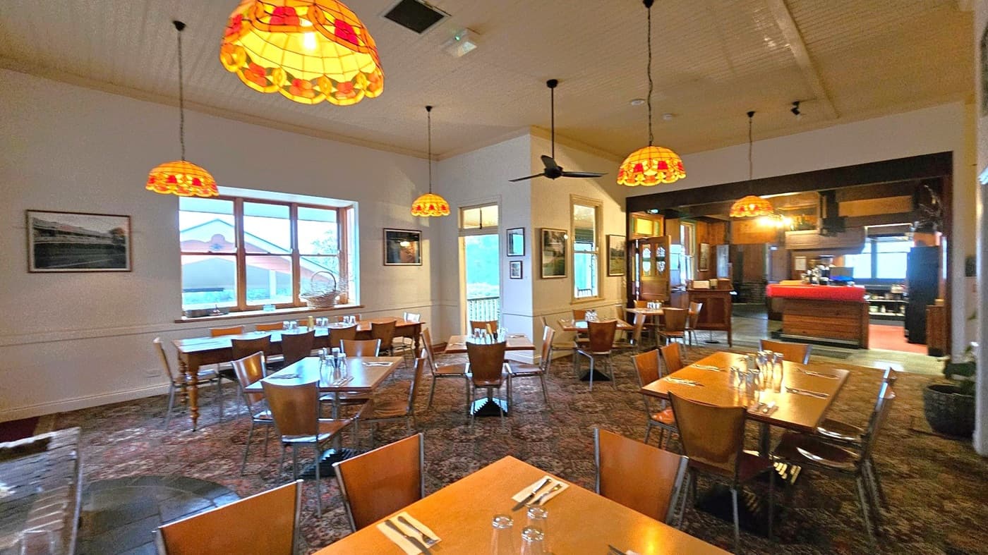 A cosy restaurant interior with wooden tables and chairs arranged neatly. The ceiling features several stained glass pendant lights with warm tones. On the left, a large window with a view of an outdoor area and framed pictures on the walls. The right side shows a bar area with a red counter and more windows. The floor is covered with a patterned carpet, and there are ceiling fans. The atmosphere is warm and inviting.