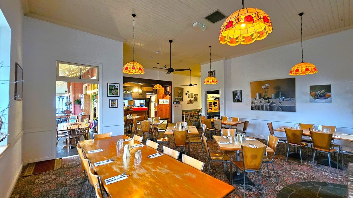 A cosy restaurant interior with wooden tables and chairs arranged neatly. The ceiling features several stained glass pendant lights with warm tones. The walls are adorned with framed artwork. In the background, there is a bar area with additional seating. The floor is covered with a patterned carpet, adding to the warm atmosphere. A doorway on the left leads to another dining area with fairy lights and more tables.