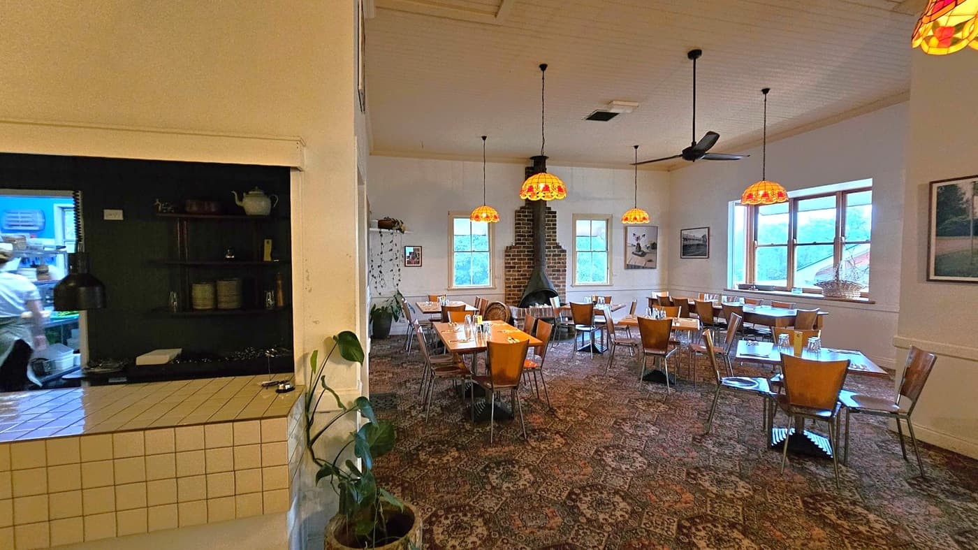 A cosy restaurant interior with a patterned carpet and wooden tables and chairs. On the left, there's a tiled counter with a view into the kitchen where a person is working. The dining area features several tables set with glasses and cutlery. Hanging from the ceiling are warm-toned pendant lights. A brick fireplace is centred on the back wall, flanked by two windows. A large window on the right lets in natural light, and there are framed pictures on the walls. A potted plant is near the counter
