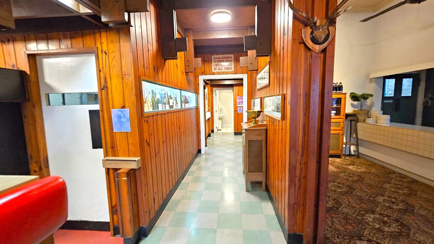 A narrow hallway with wooden panelled walls and a light-coloured tiled floor. On the left, there's a red booth seat and a small window displaying bottles. A sign above reads "CHILDREN TO REMAIN SEATED & UNDER CONTROL AT ALL TIMES." On the right, a mounted deer head and a wooden cabinet with a decorative bowl. Further right, a room with a patterned carpet, a wooden cabinet, and a table with stacked plates. A ceiling light is visible above.
