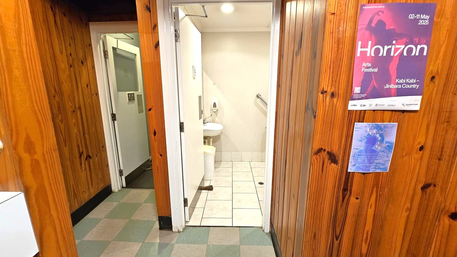 Wood-panelled walls with two posters on the right. One poster advertises the "Horizon Arts Festival" from 2-11 May 2025. A doorway in the centre leads to a small bathroom with white tiles, a sink, and a grab bar. The floor outside the bathroom is covered with green and grey checkered tiles. The ceiling is white with recessed lighting.