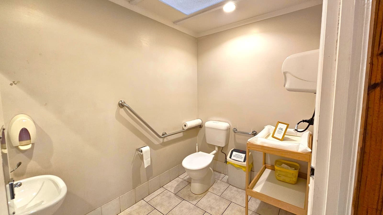 A small bathroom with light-coloured walls and tiled flooring. On the left, a sink with a soap dispenser is mounted on the wall. In the centre, a toilet with a roll of toilet paper on a metal bar is visible. On the right, a wooden changing table with a framed sign and a basket underneath is present. A waste bin is placed beside the toilet. The ceiling has a recessed light and a skylight.