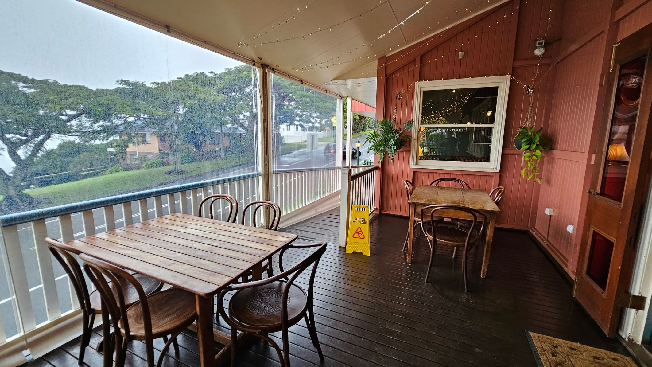 A covered porch with wooden flooring and a ceiling adorned with string lights. On the left, a wooden table with four chairs is positioned near a railing, with a view of trees and a road through a clear plastic screen. On the right, another table with chairs is set against a red wooden wall, which features a window and a hanging plant. A yellow "Caution Wet Floor" sign is placed near the centre. The atmosphere is cosy and inviting.