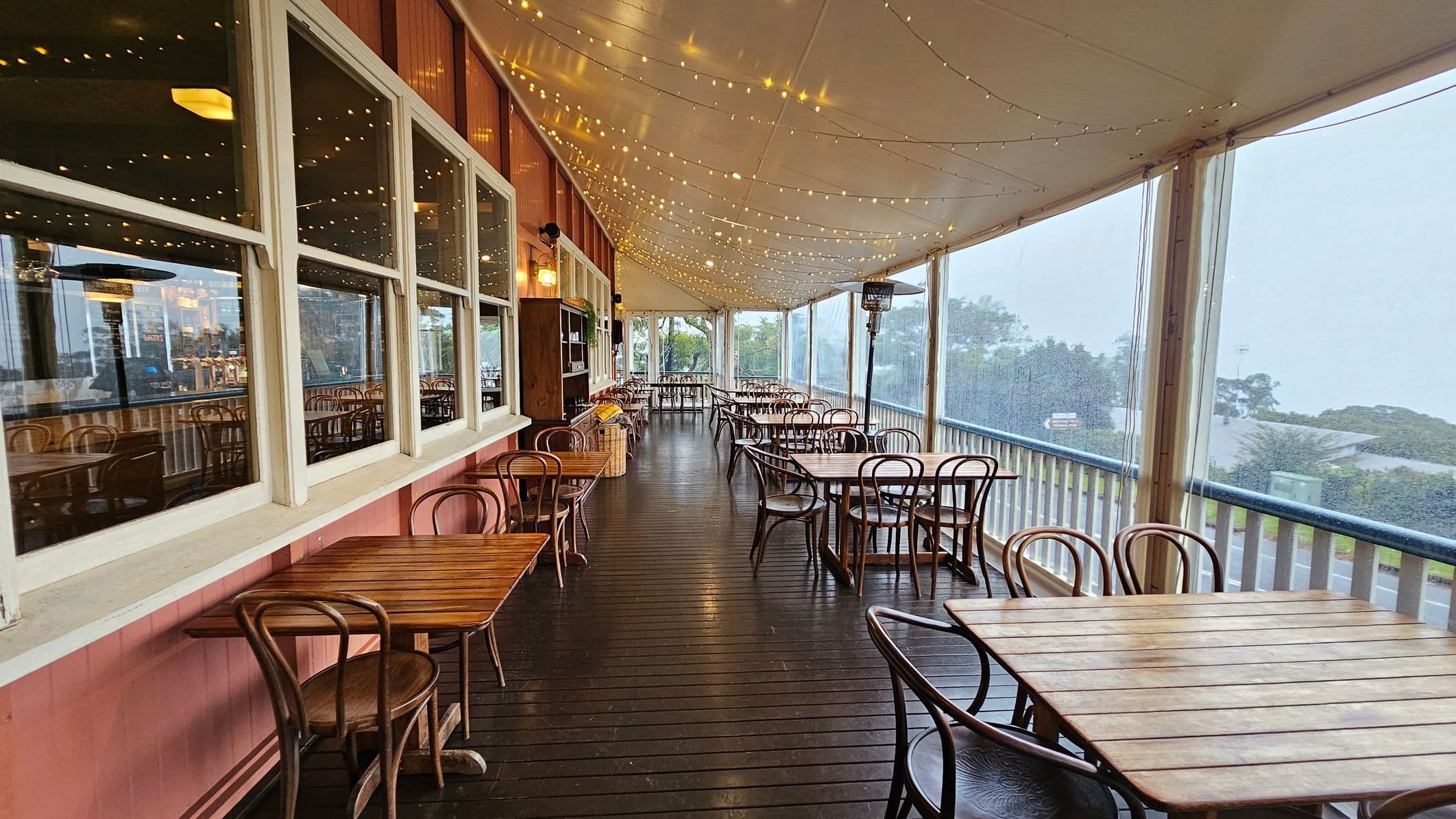 A covered outdoor dining area with wooden tables and chairs arranged along a dark wooden floor. On the left, large windows reflect the interior lights, and a shelf is mounted on the wall. The ceiling is adorned with string lights, creating a warm atmosphere. On the right, clear plastic panels offer a view of trees and a misty landscape beyond a railing. A patio heater stands among the tables.