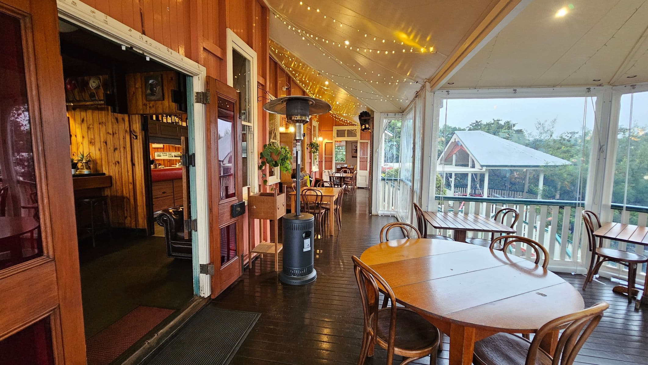 A wooden veranda with a warm, inviting atmosphere. On the left, an open door leads to an interior with wooden walls and a bar area. The right side features several wooden tables and chairs arranged along the railing, offering views of a nearby building with a green roof. The ceiling is adorned with string lights, and a tall heater stands in the centre. The floor is made of dark polished wood, enhancing the cosy feel of the space.