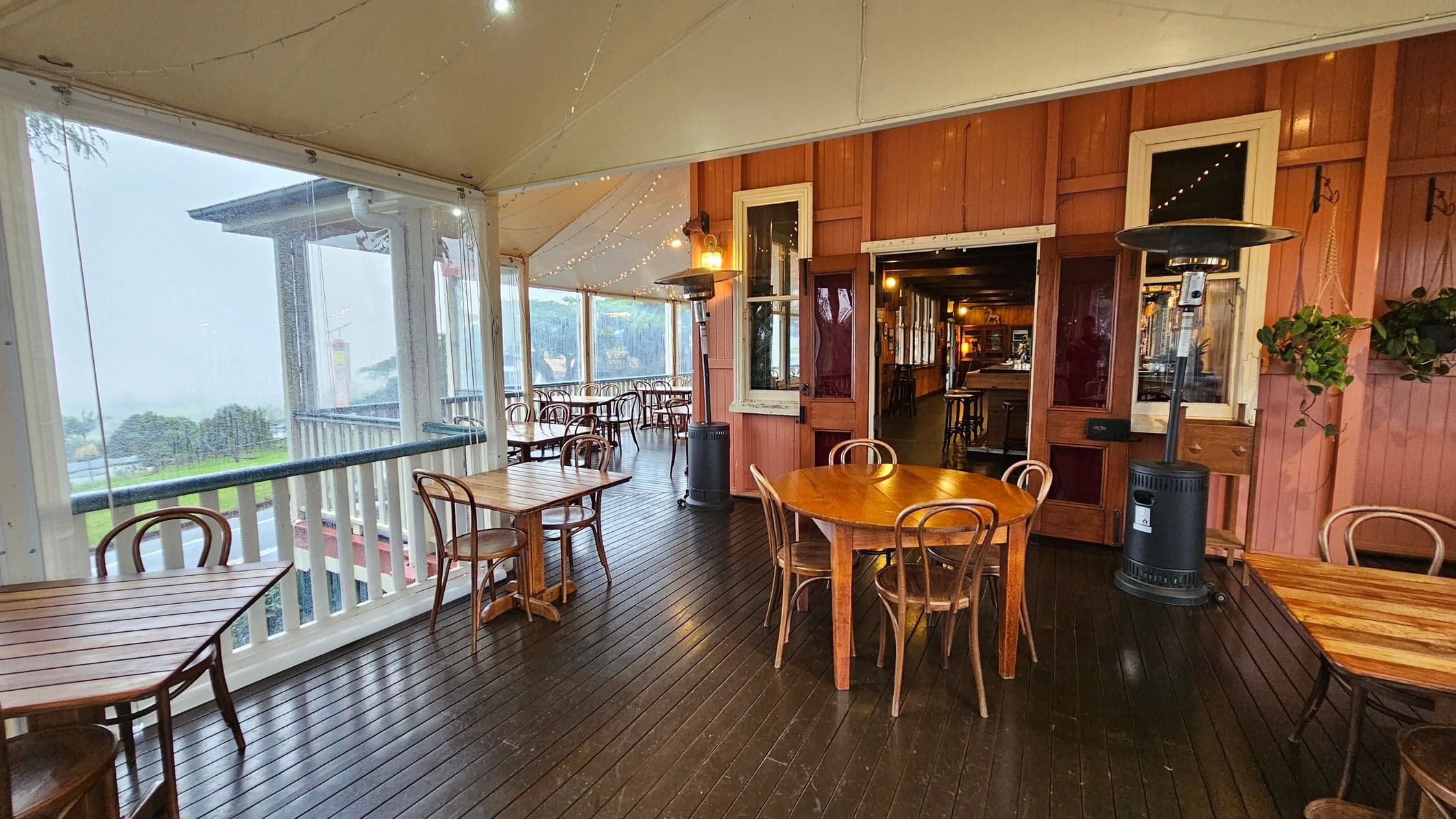 A covered outdoor seating area with wooden flooring and a light-coloured ceiling. On the left, there are wooden tables and chairs near large windows with a view of greenery and a road outside. In the centre, a round wooden table with chairs is positioned near a patio heater. On the right, the wall is wooden with windows and a door leading inside, where more seating is visible. String lights hang from the ceiling, and a potted plant is on the wall.
