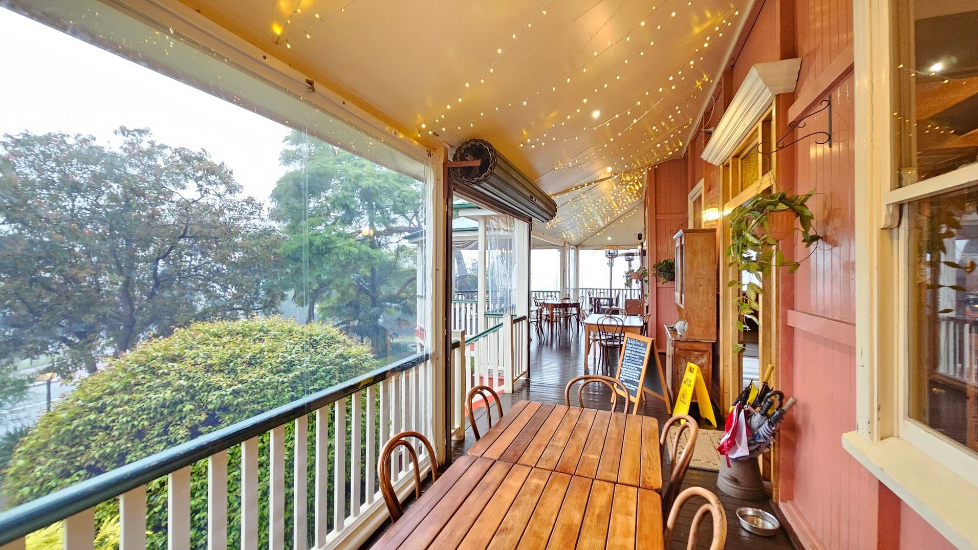 A covered veranda with wooden flooring and a ceiling adorned with string lights. On the right, a red wooden wall features windows and a door with a plant hanging nearby. A wooden table with chairs is in the foreground. On the left, clear plastic panels offer a view of lush greenery and trees outside. A small chalkboard sign and a container with umbrellas are near the entrance.