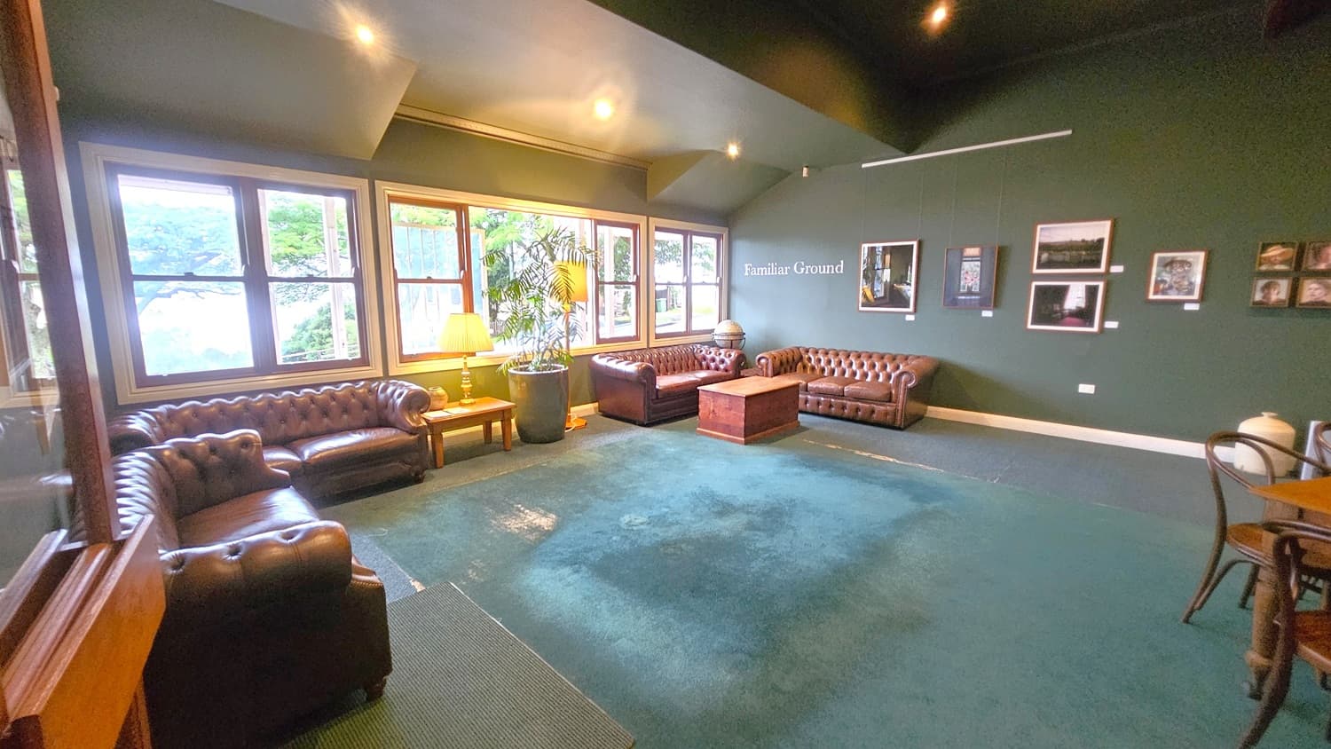 A room with green walls and a green carpeted floor. On the left, there are large windows with a view of greenery outside, and a brown leather sofa beneath them. A lamp and a potted plant are next to the sofa. In the centre, another brown leather sofa and a wooden coffee table are visible. The right wall displays framed pictures and the words "Familiar Ground." A wooden chair and table are partially visible in the foreground on the right. The ceiling is angled with recessed lighting.