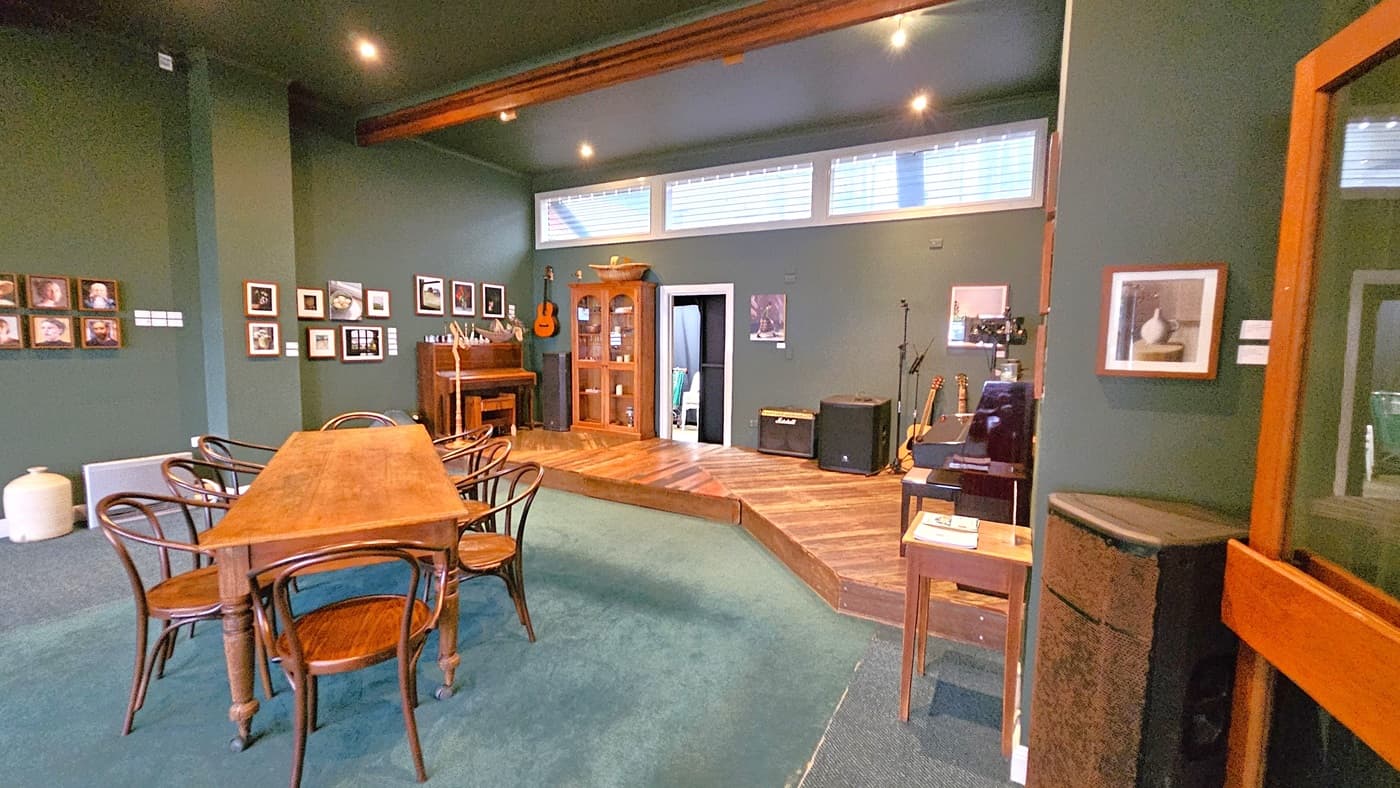 A room with dark green walls and a wooden floor. On the left, there's a wooden table surrounded by chairs, and framed pictures on the wall. In the centre, a small stage area with a piano, a guitar hanging on the wall, and a cabinet. On the right, musical equipment including an amplifier and guitars. The ceiling has recessed lighting, and there are high windows letting in natural light. A small table with a book is near the foreground.