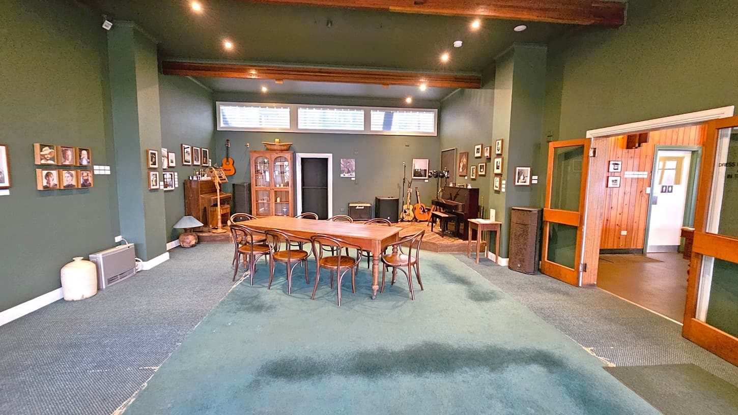 A room with green walls and a green carpeted floor. In the centre, a long wooden table with several chairs. The left wall displays framed photos and a small heater. The back wall has a glass cabinet, guitars, and a piano. The right side features a wooden door leading to another room with wood-panelled walls. The ceiling has exposed wooden beams and recessed lighting.