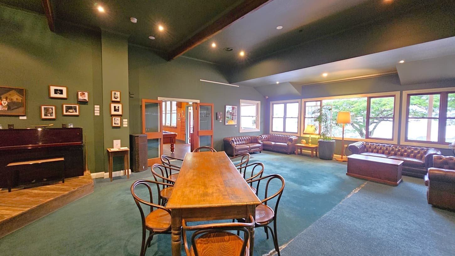 A spacious room with green walls and a green carpet. On the left, a dark piano is positioned against the wall, surrounded by framed pictures. In the centre, a long wooden table with several chairs is placed on the carpet. The right side features large windows with views of trees outside, and several brown leather sofas with a coffee table. A floor lamp and a potted plant are near the windows. The ceiling has recessed lighting, and a doorway leads to another room with a pool table.