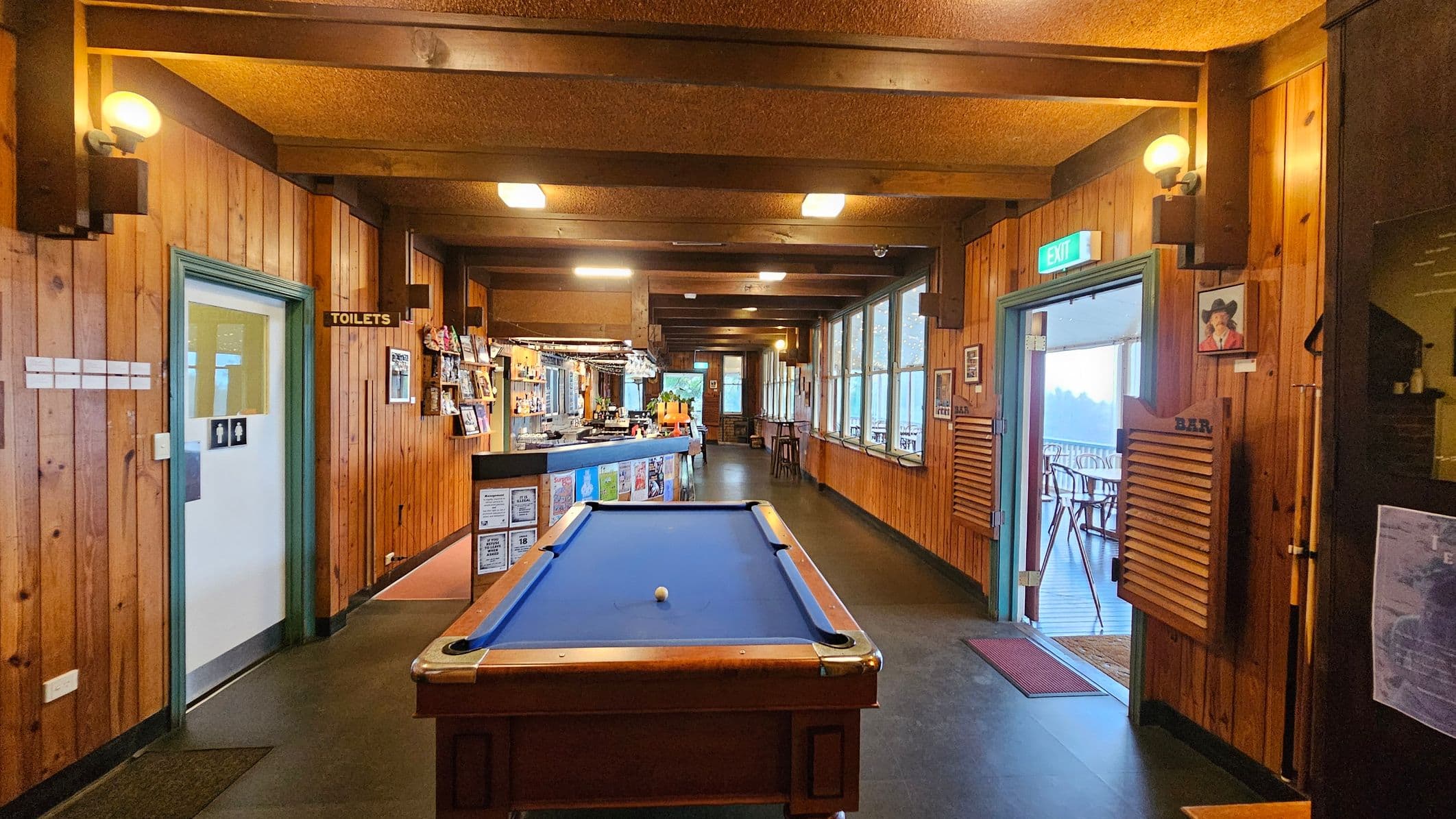 A cosy bar interior with wooden walls and ceiling beams. In the centre, a blue pool table with a single white ball. To the left, a door marked "Toilets" with a sign above. On the right, a door with a green "Exit" sign leads to an outdoor seating area. The bar counter is visible in the background, adorned with various items and surrounded by stools. Large windows line the right wall, allowing natural light to illuminate the space.