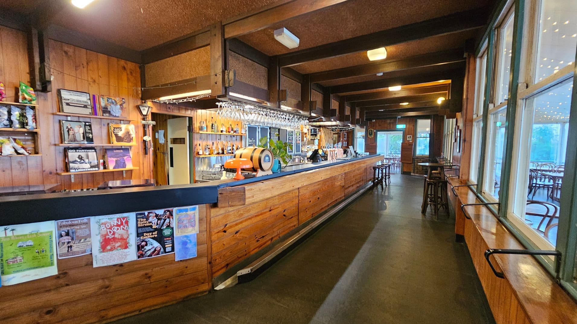 A cosy bar interior with wooden walls and ceiling. On the left, shelves display vinyl records and snacks. The long wooden bar counter features posters on the front and is topped with glasses, bottles, and a small barrel. Hanging lights illuminate the area. On the right, large windows with railings overlook a seating area with round tables and chairs. The floor is dark, and the ceiling has exposed beams with lights.