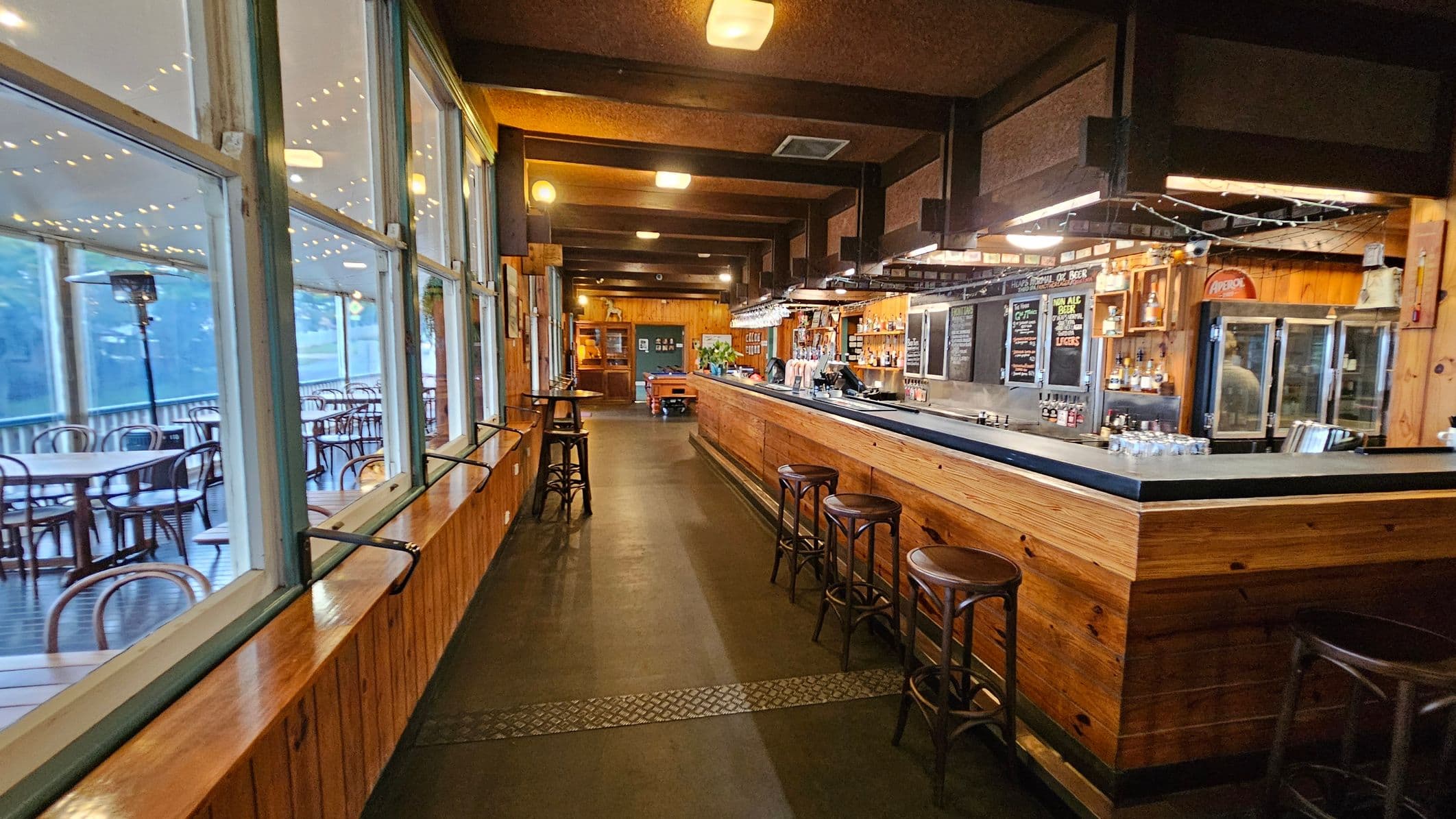 A cosy bar interior with a long wooden counter on the right, lined with high stools. Behind the bar, shelves display various bottles and chalkboards list menu items. On the left, large windows reveal an outdoor seating area with round tables and chairs. The ceiling is wooden with exposed beams, and the floor is dark with a patterned strip running down the centre. Warm lighting creates an inviting atmosphere.